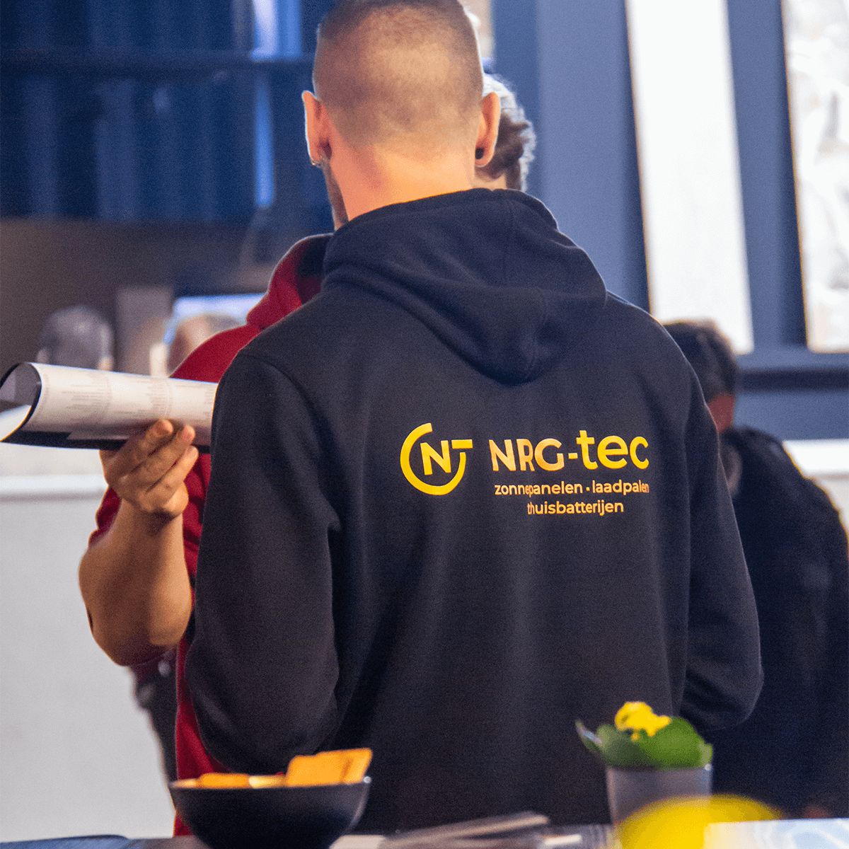 Medewerker Gilles van NRG-TEC in zwarte hoodie met bedrijfslogo terwijl hij documentatie en informatie deelt op een bouwbeurs in Roeselare over zonnepanelen, laadpalen en thuisbatterijen.